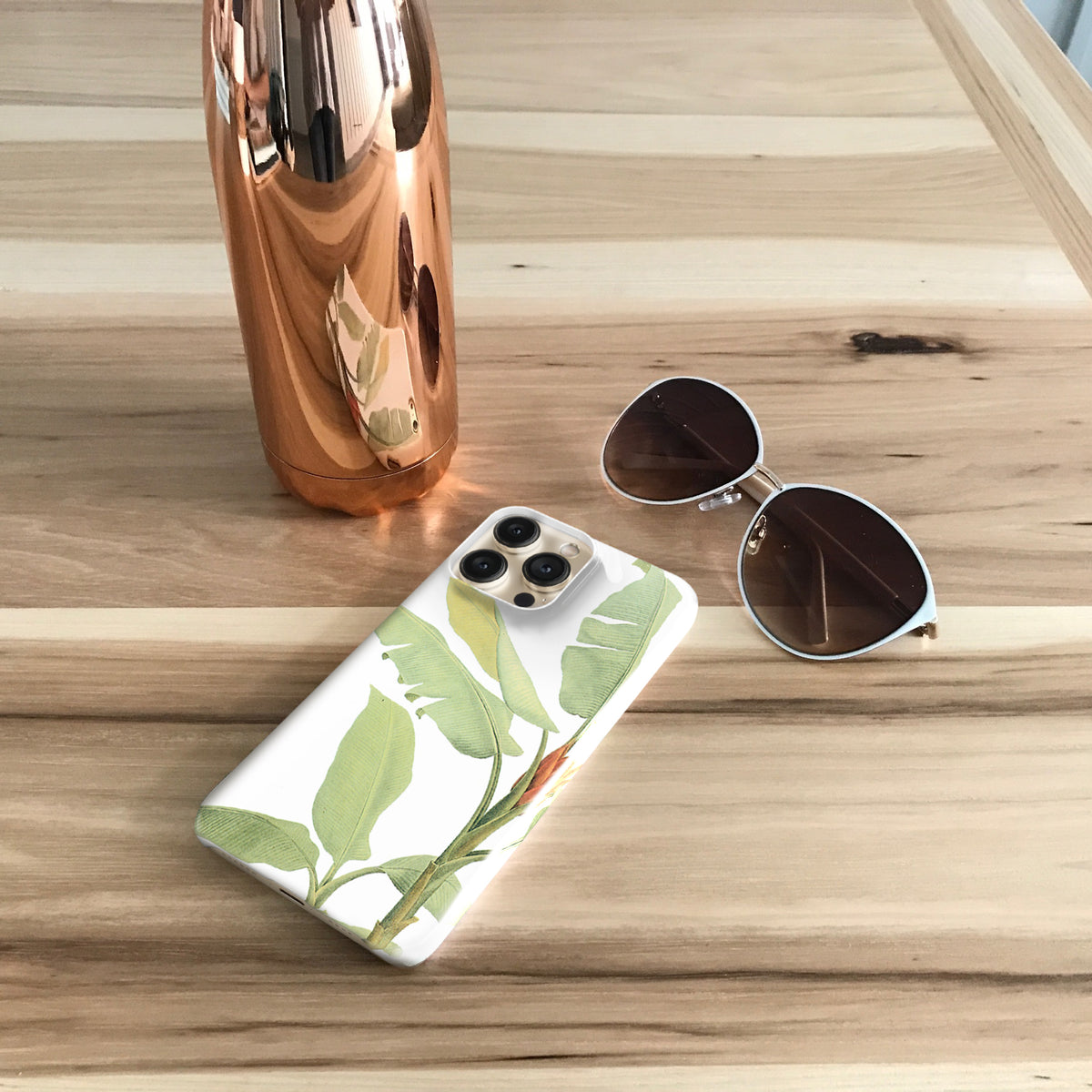 A white phone case with a green leaf pattern, a pair of sunglasses, and a copper-colored water bottle are arranged on a wooden surface.