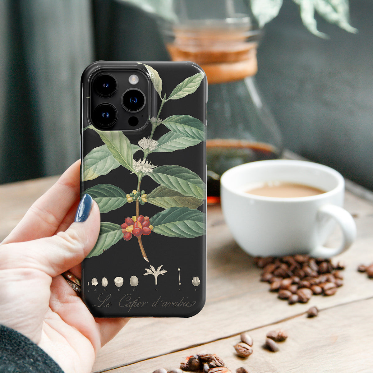 A person&#39;s hand holding a phone with a floral design on the back cover, with a cup of coffee and coffee beans on a table in the background.