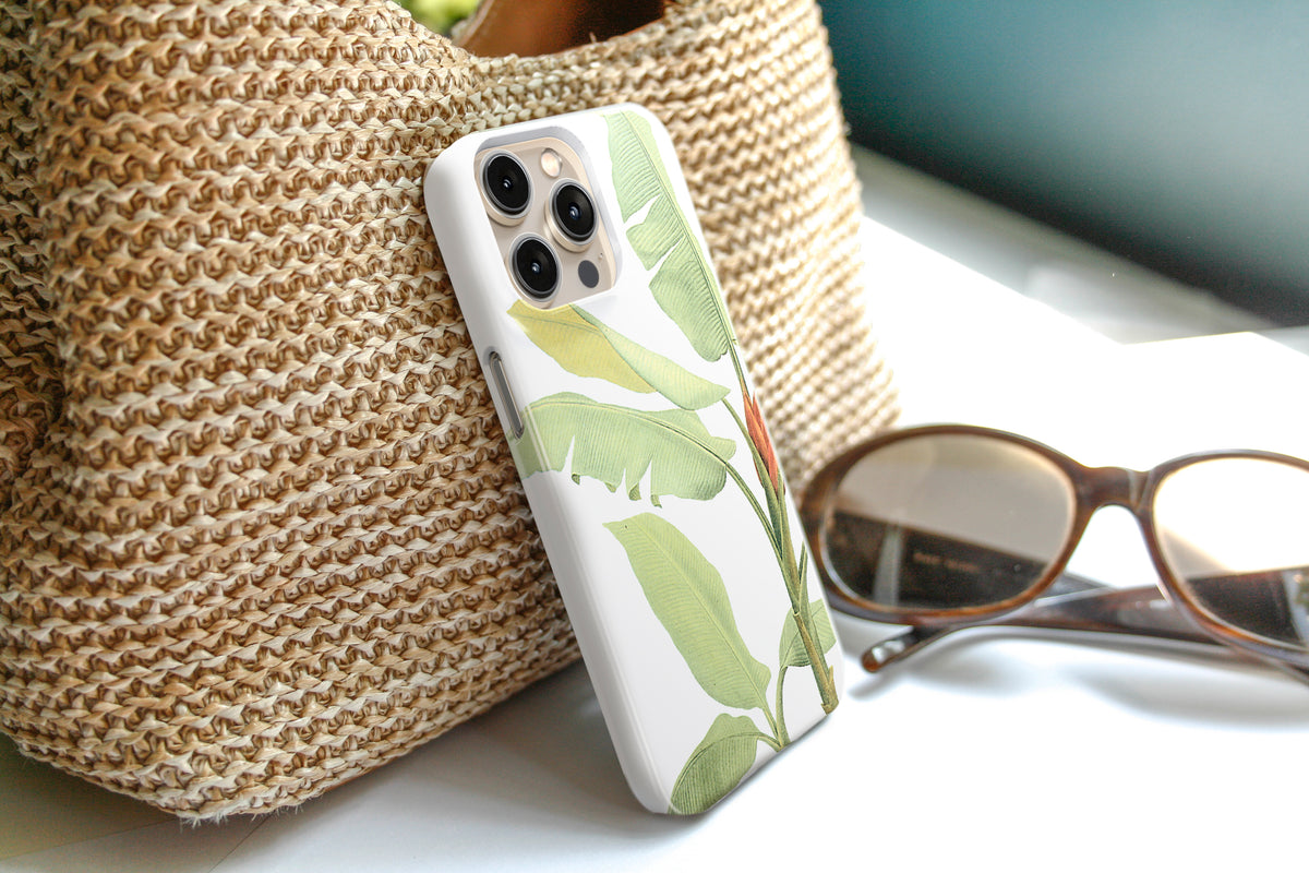 A white phone case with a green leaf pattern is placed on a woven straw bag, with a pair of sunglasses and a pair of glasses resting on a white surface nearby.