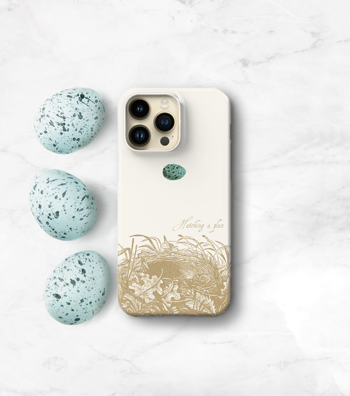 A white iPhone case with a gold design depicting a bird&#39;s nest and three speckled eggs is placed on a marble surface.